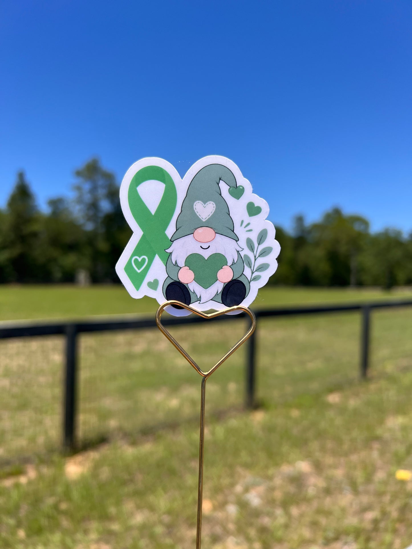 Sticker of a gnome with a green ribbon on a metal hook against a blue sky and grass background.