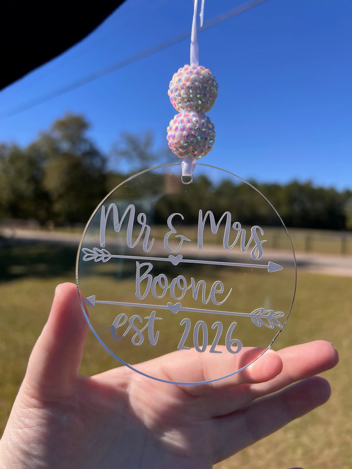 A hand holding a clear round acrylic wedding ornament with personalized text reading 'Mr & Mrs Boone est 2026' and arrow details, alongside two iridescent beaded strands against an outdoor background.