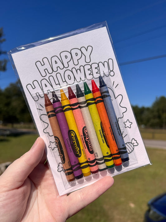 A hand holds a clear plastic package containing eight Crayola crayons attached to a white card that says 'HAPPY HALLOWEEN!' in large outlined letters. The background shows an outdoor scene with blue sky, trees, and grass.