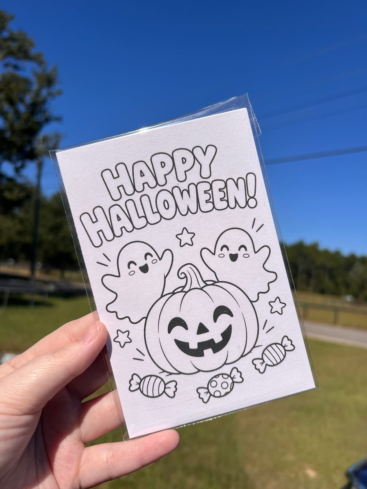 A hand holds a clear-sided card featuring a black-and-white Halloween illustration with the words “HAPPY HALLOWEEN!” and a jack-o'-lantern, ghost faces, stars, and candy, against an outdoor background with grass, trees, and blue sky.