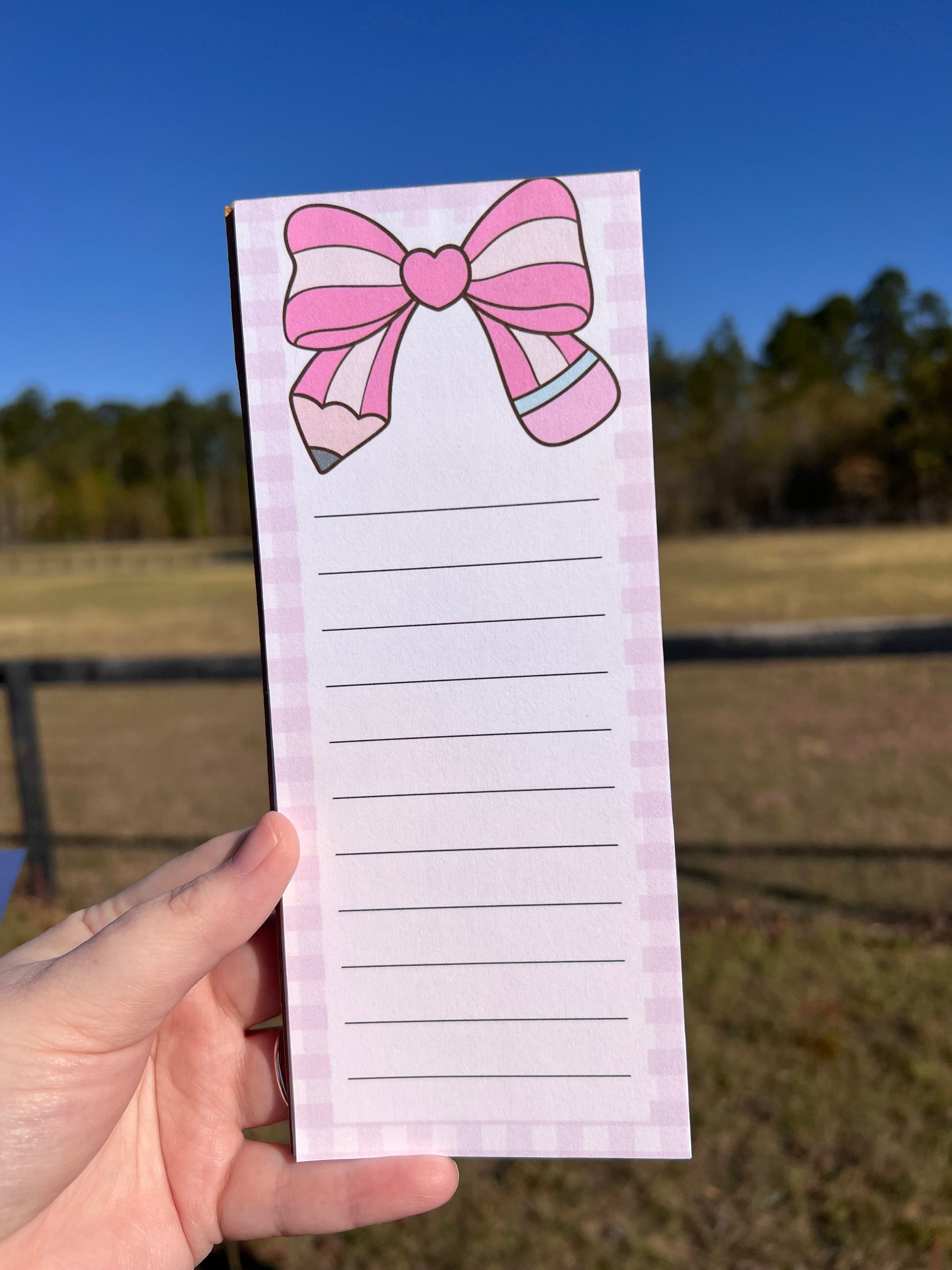 Notepad with a pink bow design held by a hand against a blurred outdoor background