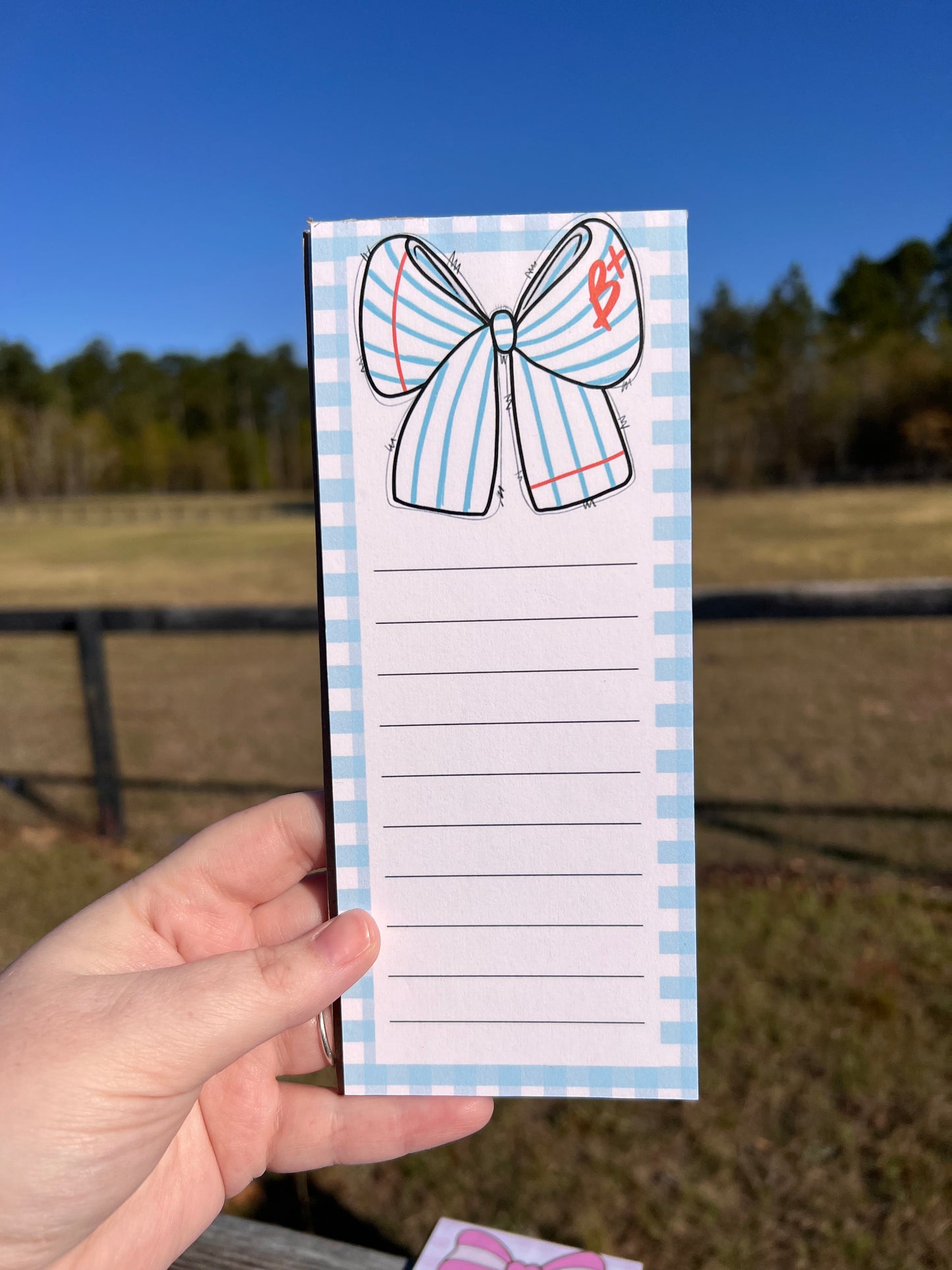 Hand holding a notepad with a decorative bow design against a blurred outdoor background