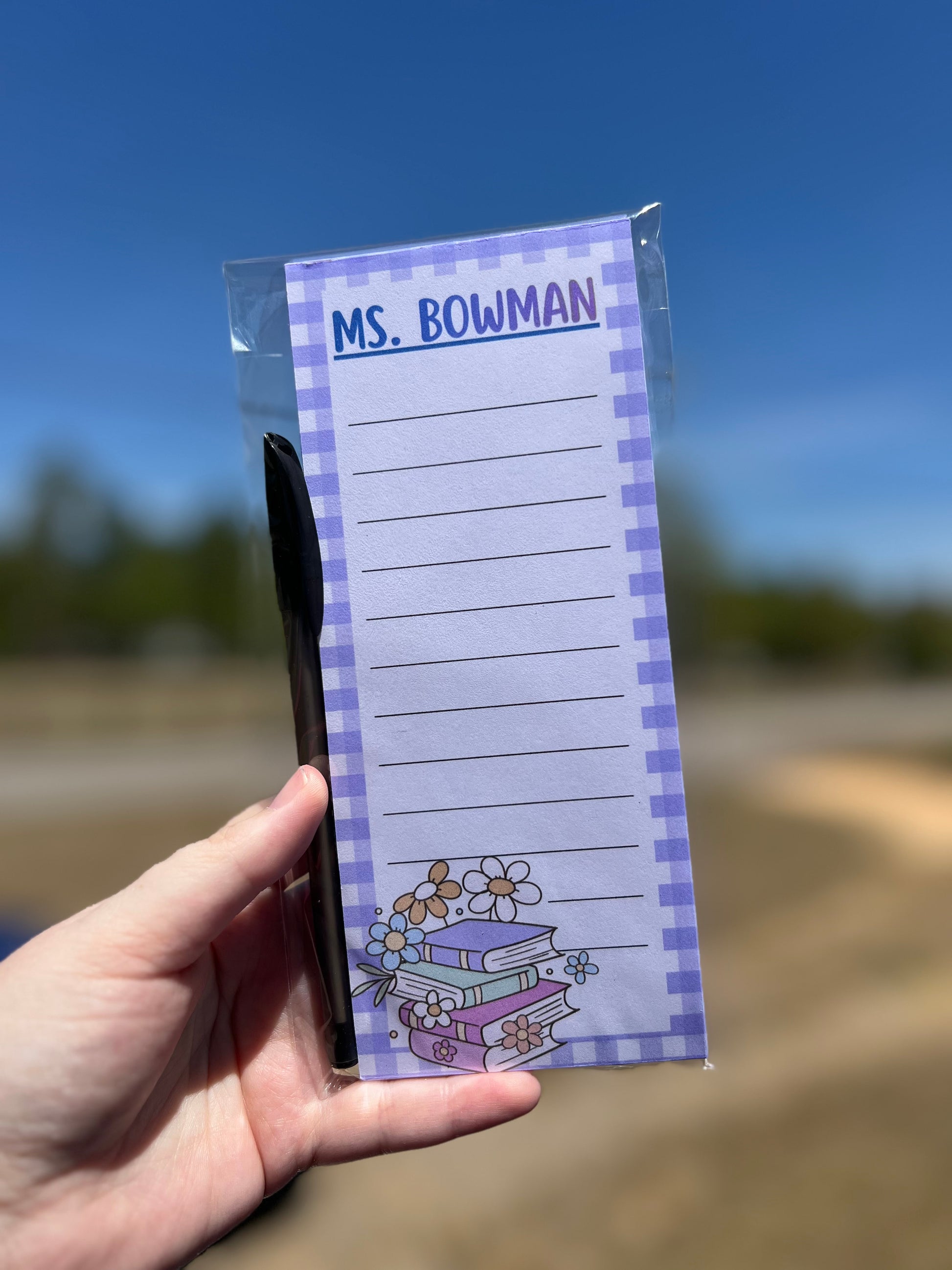 Notepad with 'MS. BOWMAN' printed on it held by a hand against a blurred outdoor background.