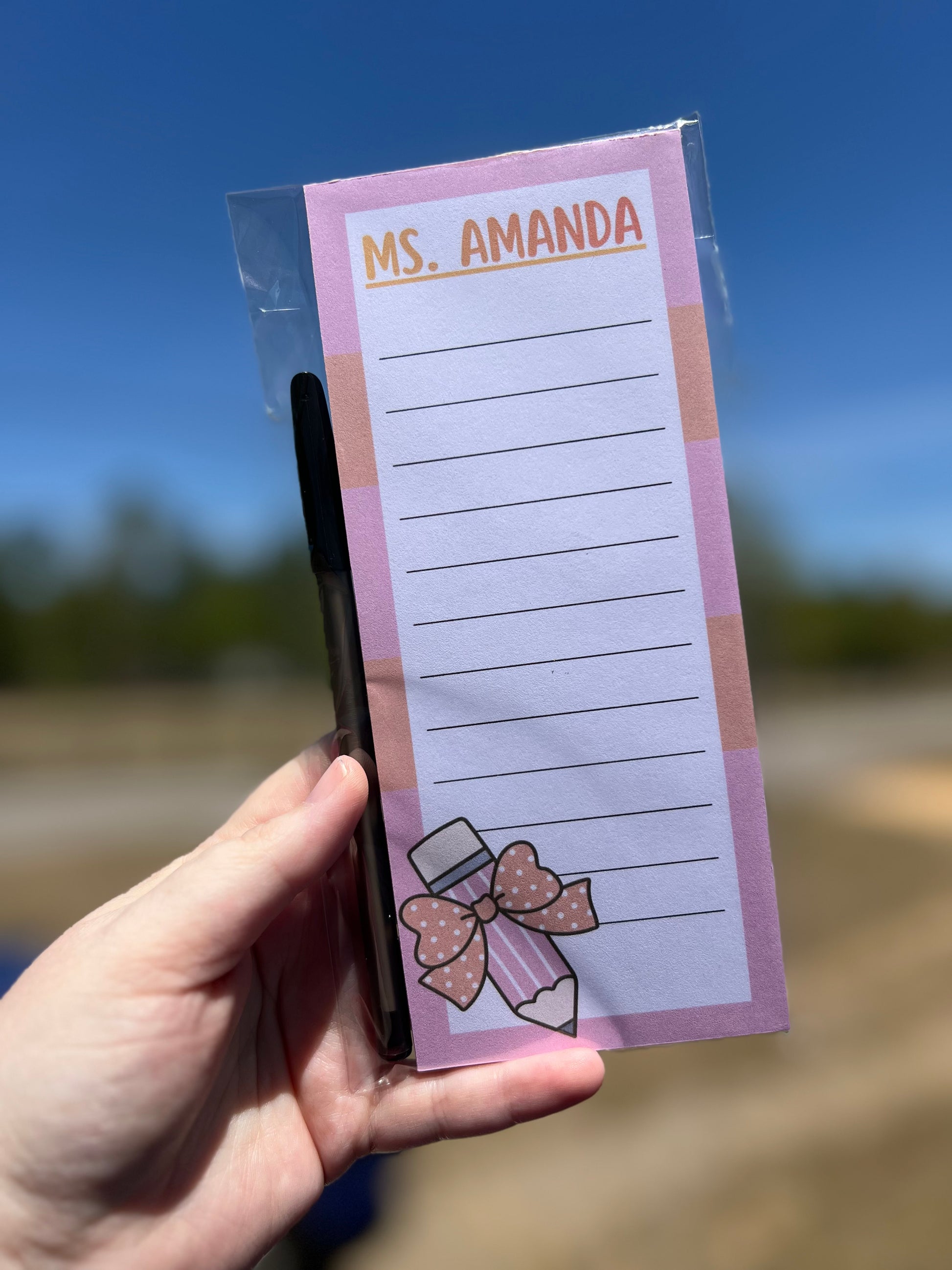 Notepad with pen holder labeled 'Ms. Amanda' held by a hand against a blurred outdoor background