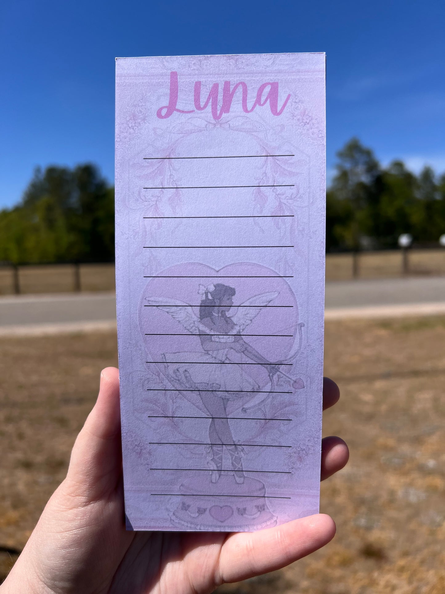 Hand holding a pink notebook with 'Luna' and a design on the cover against a blue sky.