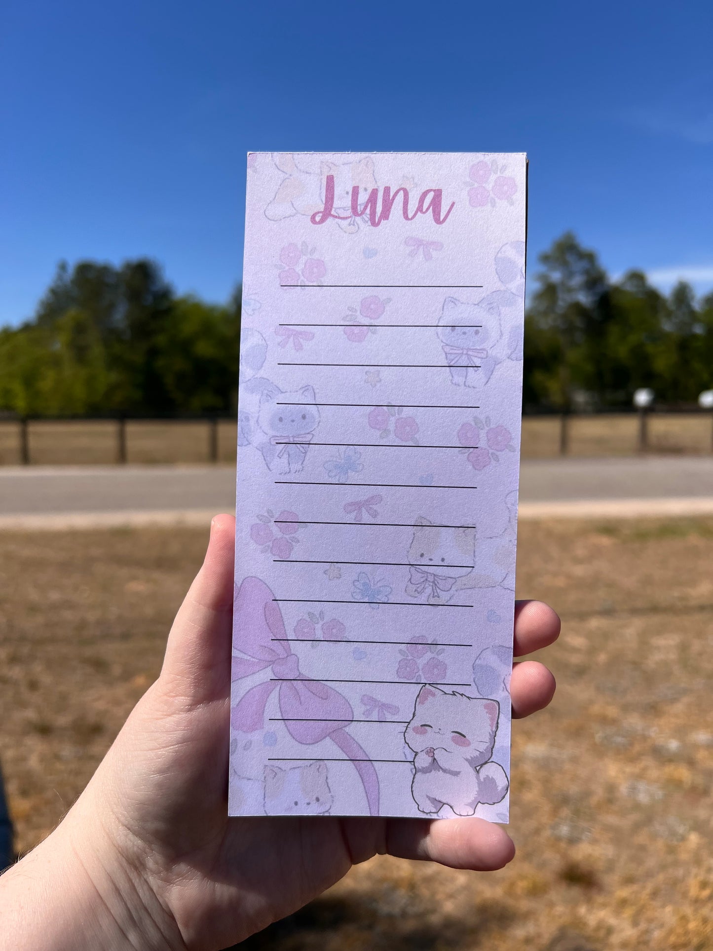 Hand holding a notepad with a bear design and 'Luna' printed on it against a blue sky.