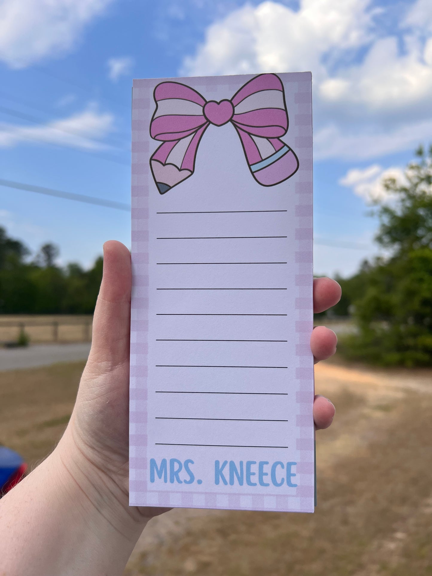 Notepad with a pink bow design held by a hand against a blurred outdoor background