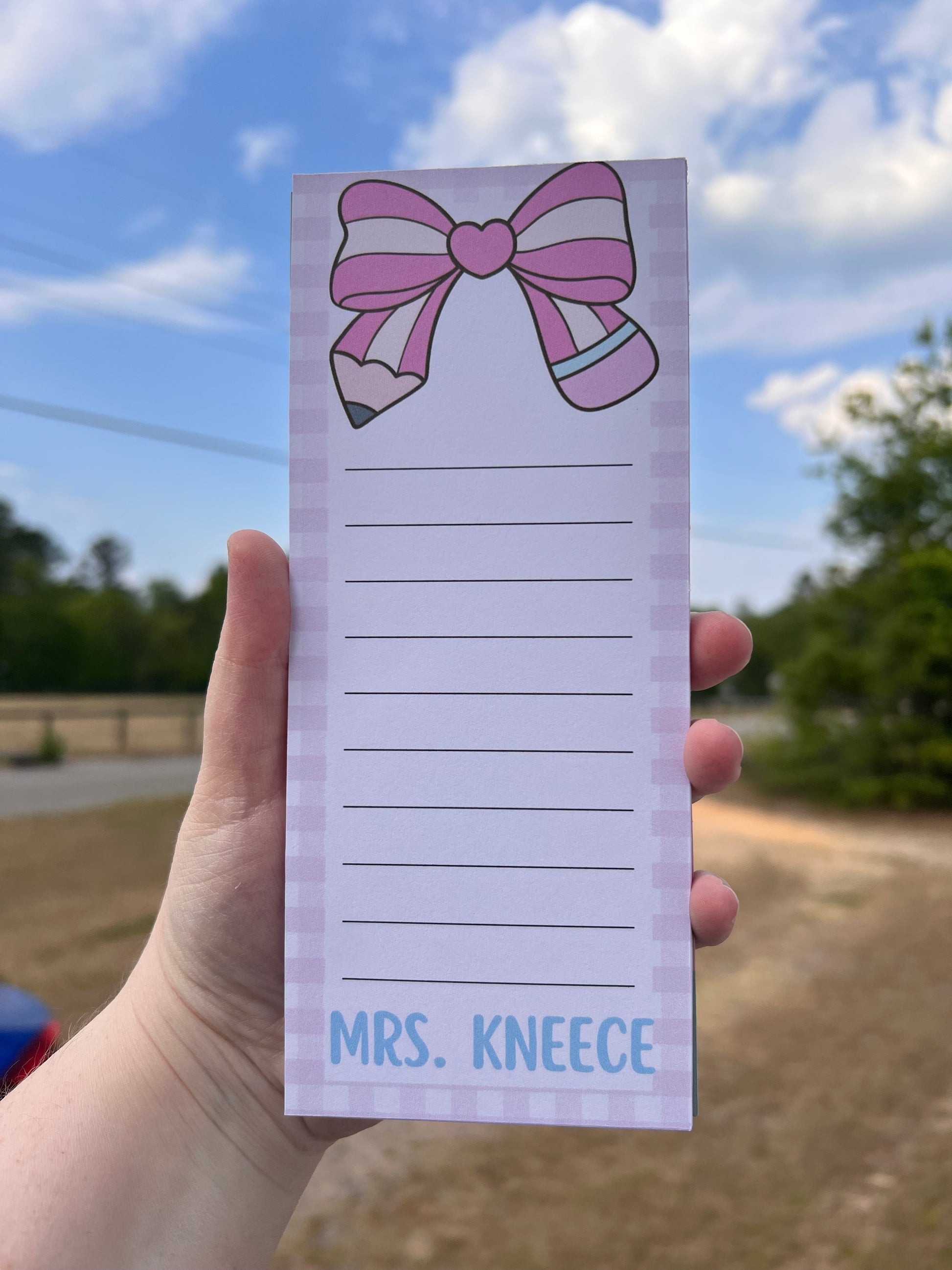 Notepad with a pink bow design held by a hand against a blurred outdoor background
