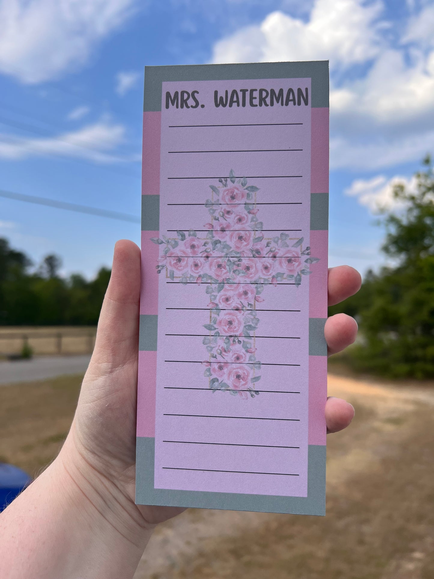 Hand holding a floral notepad with 'Mrs. Waterman' printed on it against a blurred outdoor background.