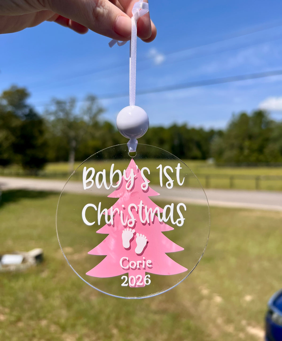 A hand holds a clear round hanging ornament outdoors. The ornament has a pink Christmas tree silhouette with white script text reading 'Baby's 1st Christmas' and small white baby footprints, plus the name 'Corie' and year '2026'.