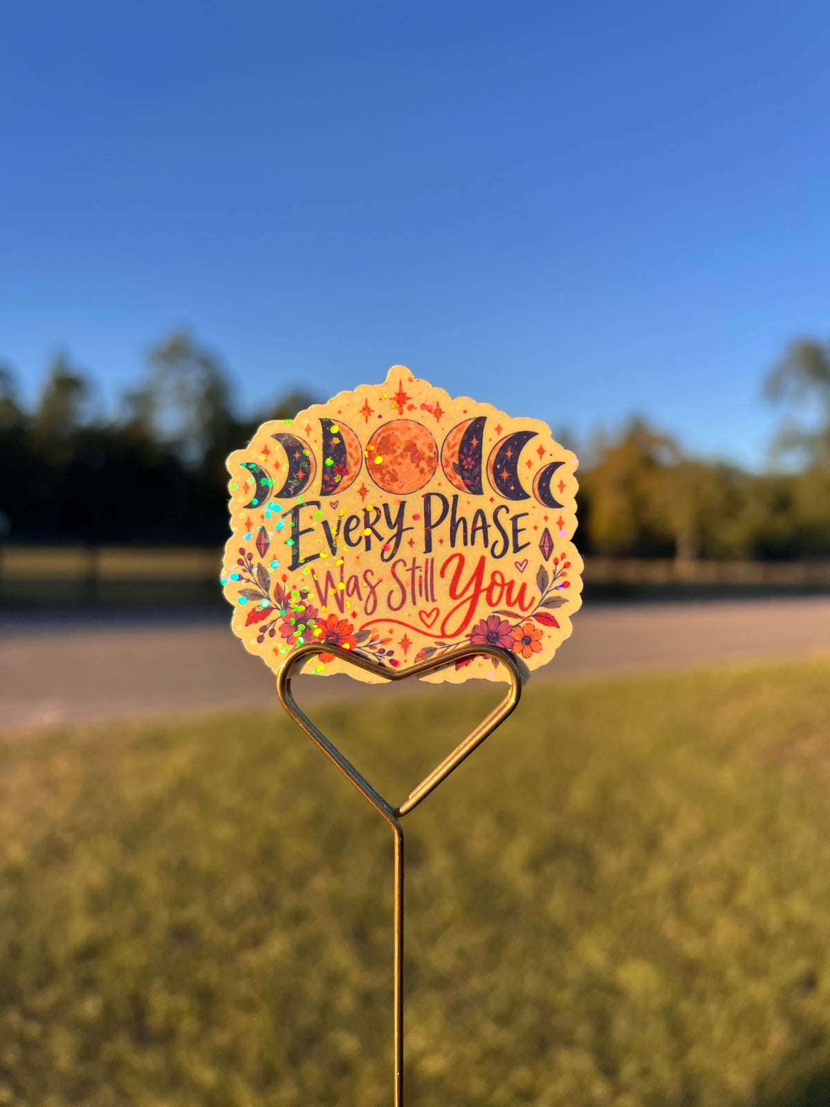 A decorative die-cut sign with a moon phase design and the phrase 'Every Phase Was Still You' in colorful lettering, mounted on a heart-shaped metal stake outdoors against a blurred grassy background and blue sky.