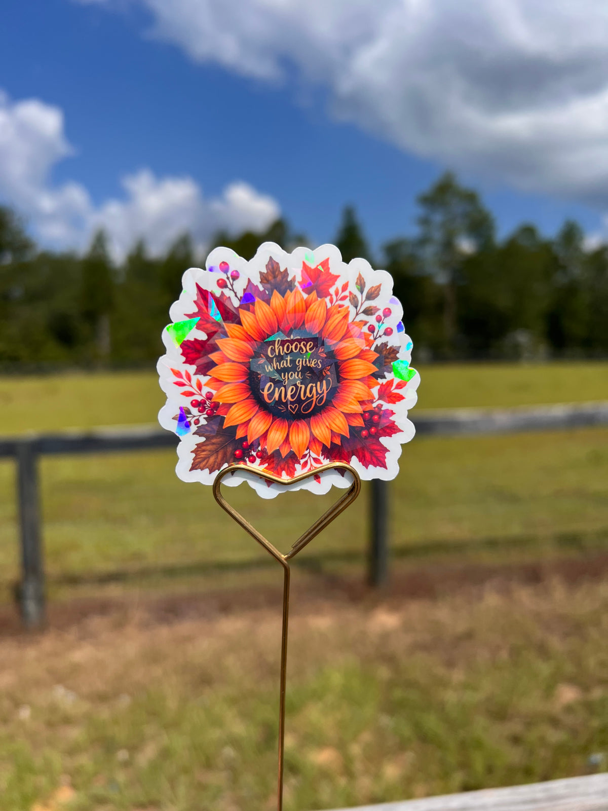 A die-cut sticker featuring a colorful autumn sunflower and fall leaves with the text 'choose what gives you energy' in script, displayed on a metal stake in an outdoor grassy field with a fence and blue sky in the background.